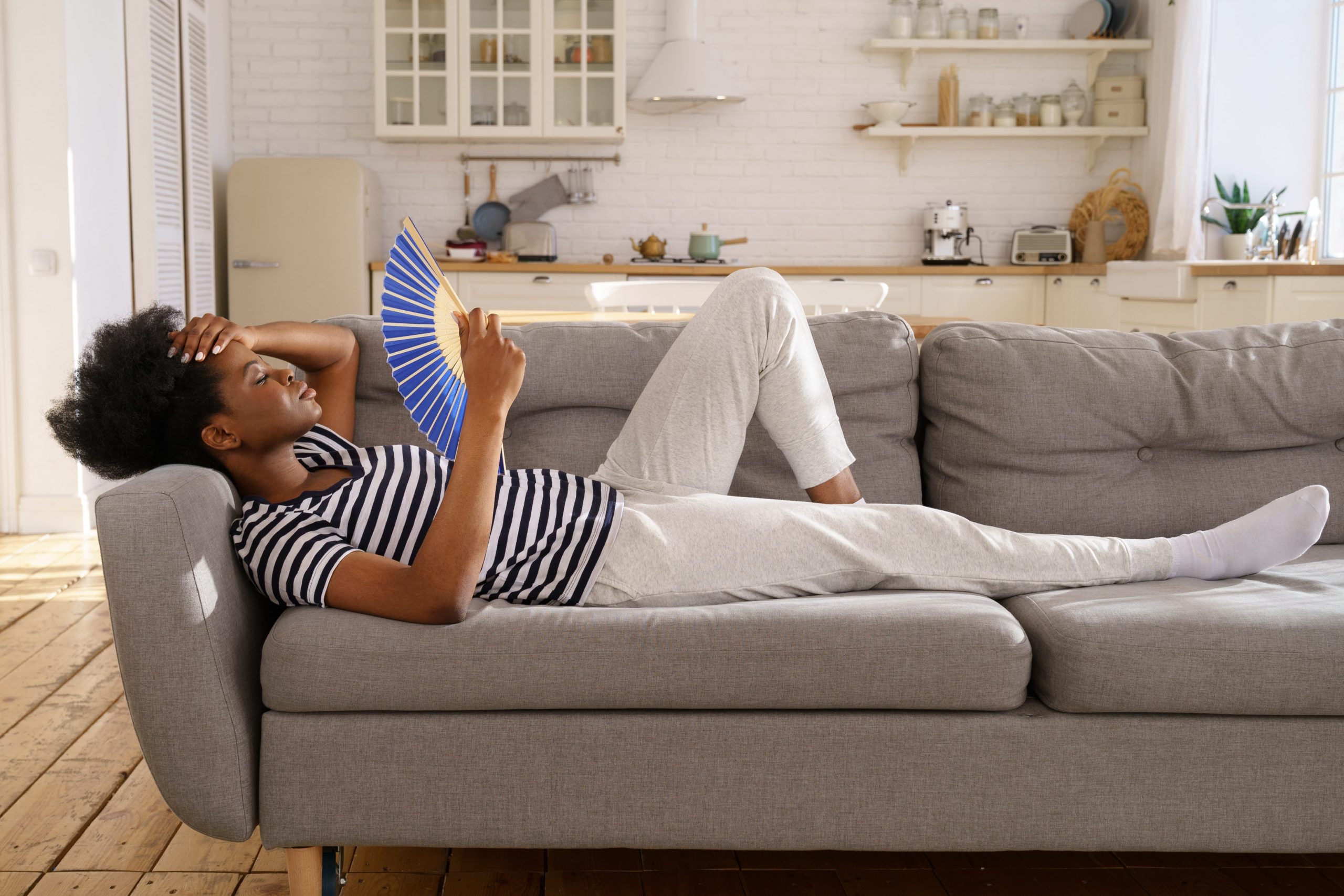 young woman on couch in apartment trying to cool off with fan